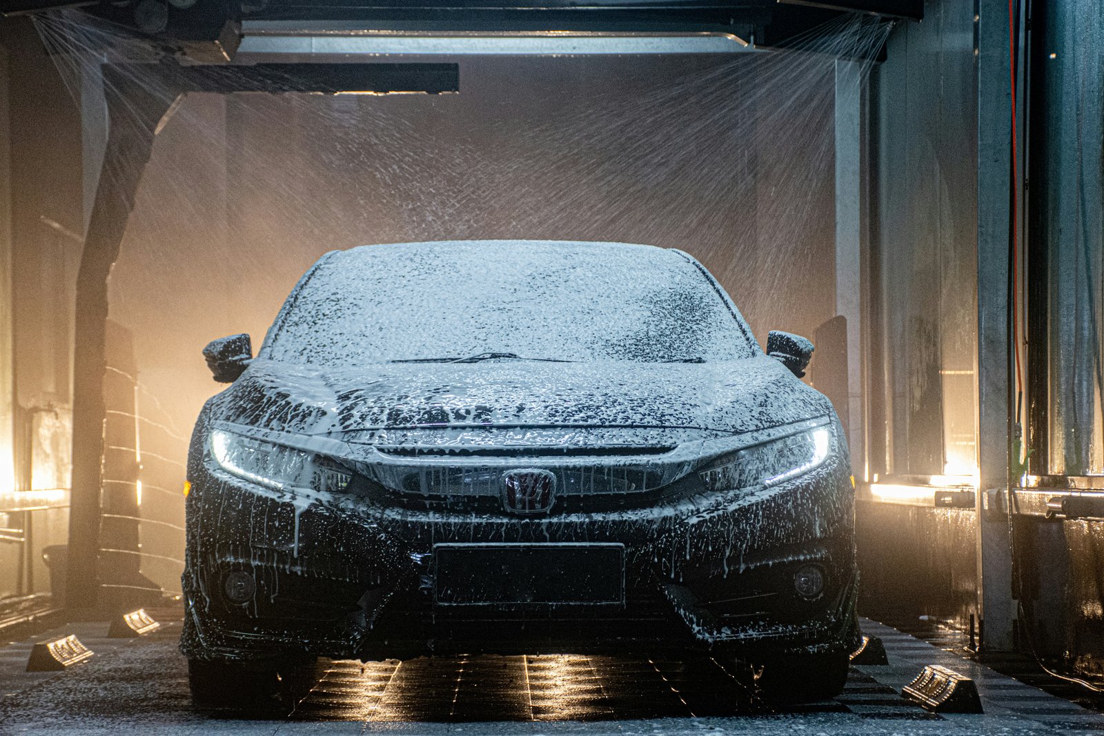 A dark sedan covered in foam inside an automatic car wash, lit from behind